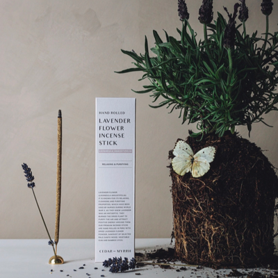 Product photo of the Lavender Flower Incense Stick. A box of incense standing upright on a table with an uprooted lavender plant sitting beside it with a butterfly resting on the plant.