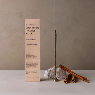 Product photo of the Cinnamon Incense Stick. A box of incense sitting upright on a table with a lit incense stick beside it and cinnamon sticks arranged on the table.